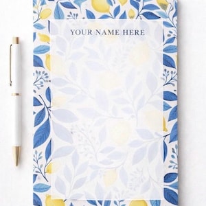 Op de afbeelding: Notitieblok met een citroen- en bladerenontwerp, met blauwe bladeren en gele citroenen. Het notitieblok heeft een witte achtergrond met de tekst "YOUR NAME HERE" bovenaan. Een witte pen met gouden accenten ligt naast het notitieblok.