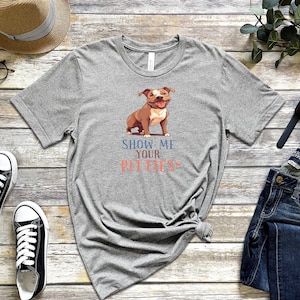 Pode incluir: Camiseta cinza com uma imagem de desenho animado de um pit bull marrom e branco. O texto "Show me your pitties" está impresso na camiseta.