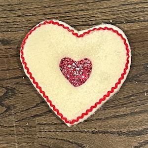 Könnte beinhalten: Ein weißes Filzherz mit rotem Filzrand und einem kleineren pinkfarbenen Glitzerherz in der Mitte.
