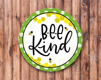 Op de afbeelding: Een rond, groen en wit bord met een honingraatpatroon en witte madeliefjes rond de rand. Het bord zegt "bee kind" in zwarte letters met een bij-illustratie aan elke kant van de tekst.
