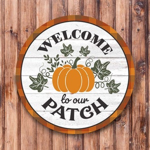Könnte beinhalten: Rundes Holzschild mit weißem Hintergrund und orange-schwarzem Karomuster am Rand. Das Schild trägt die Aufschrift "Willkommen in unserem Patch" mit einem Kürbis und Blättern in der Mitte.