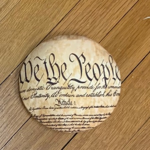 Puede incluir: Un posavasos redondo de color crema con el texto "We the People" y la primera parte del preámbulo de la Constitución de los Estados Unidos impreso en él.