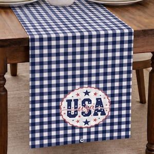 Puede incluir: Camino de mesa a cuadros azules y blancos con un diseño patriótico de EE. UU. El camino de mesa presenta un patrón de cuadros repetido y un emblema ovalado central con la palabra "USA" en rojo y azul, rodeado de estrellas.