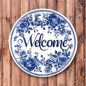 May include: Round white sign with a blue floral design and the word "Welcome" in elegant script. The sign is mounted on a wooden background, creating a welcoming aesthetic. Ideal for home decor.