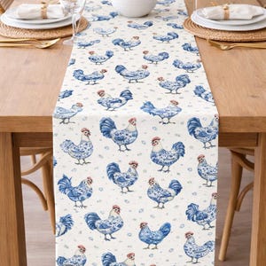 May include: A white table runner with a repeating pattern of blue roosters. The roosters have detailed blue feathers and red combs. The runner is placed on a wooden table set for a meal, with white plates and glasses.