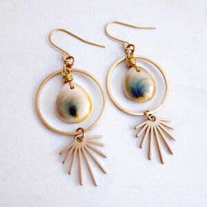 May include: A pair of gold-toned dangle earrings featuring a circular hoop frame. Each earring suspends a smooth, teardrop-shaped shell with blue and cream swirls, accented by a gold-toned sunburst charm attached to the bottom of the hoop.