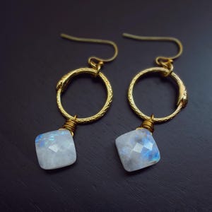 May include: A pair of gold-colored earrings with a circular design and a square, iridescent gemstone. The earrings have a hook closure and are set against a dark background. The gemstones have a blue and white color.