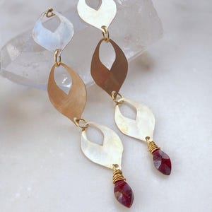 May include: A pair of long, dangling earrings. Each earring features three gold-toned, leaf-shaped pendants connected by small rings. The bottom of each earring has a faceted, deep red gemstone wrapped in gold wire. The earrings are displayed on a white surface.