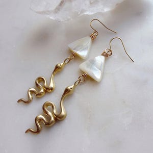 May include: A pair of gold-colored earrings featuring a snake design. Each earring has a pearlescent, triangular-shaped charm at the top, connected to a detailed snake pendant. The earrings are displayed on a white marble surface.