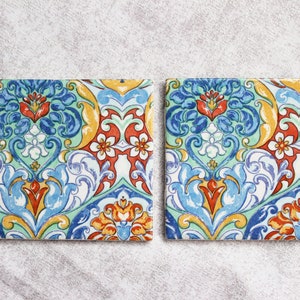 May include: Two square ceramic coasters with a colorful floral pattern. The pattern features blue, green, yellow, red, and white colors. The coasters have a glossy finish.