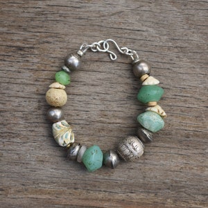 May include: A handcrafted bracelet featuring a mix of beads in various shapes and colors. The bracelet includes silver-toned metal beads, sea glass, and decorative beads with a unique clasp. The bracelet is displayed on a wooden surface.