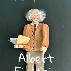 May include: A wooden ornament depicting Albert Einstein with white hair, wearing a tan jacket, brown tie, and trousers. The figure holds an open book. Text reads New for 2026 and Albert Einstein. A metal hook is attached to the top for hanging.