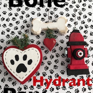 May include: A collection of dog-themed ornaments, including a bone, a heart, and a fire hydrant. The ornaments are red, white, and black, with the word "Bone", "Hydrant", and "Paw" in the image. The background is white with black paw prints.