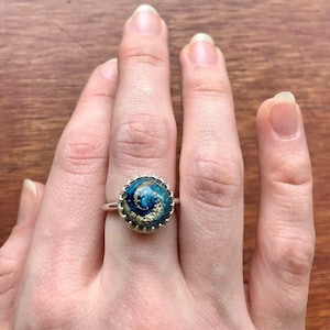 Può includere: Un anello d'argento con un design a spirale blu e oro. L'anello è incastonato in una ghiera d'argento e ha un bordo testurizzato.