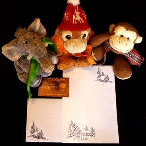 May include: Three plush toys, a gray elephant, a brown monkey, and a red and white monkey wearing a Santa hat, are arranged on a white background with a black and white drawing of a cabin in the woods. The drawing is repeated on two pieces of paper. A business card for "The Smiling Monkey Shop" is also visible.