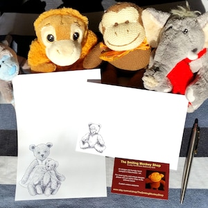 May include: A white card with a hand-drawn illustration of two teddy bears. The card is sitting on a striped blue and white surface. There are three stuffed animals in the background: a brown monkey, an orange monkey, and a gray elephant.
