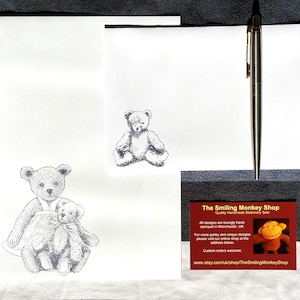 May include: A white card with a black and white illustration of a teddy bear holding a smaller teddy bear. The card has the text "The Smiling Monkey Shop" and "Quality Handmade Stationery Sets".