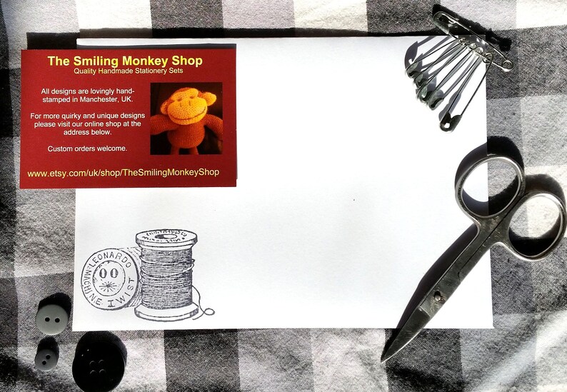 Puede incluir: Un sobre blanco con una tarjeta de visita roja de The Smiling Monkey Shop, con una ilustraci&oacute;n de un mono. Unas tijeras, imperdibles y botones est&aacute;n sobre una superficie a cuadros.