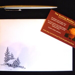 May include: A white card with a black and white illustration of a small cabin with trees. The card is next to a business card for The Smiling Monkey Shop, which features a smiling monkey illustration and the website address www.etsy.com/uk/shop/TheSmilingMonkeyShop.