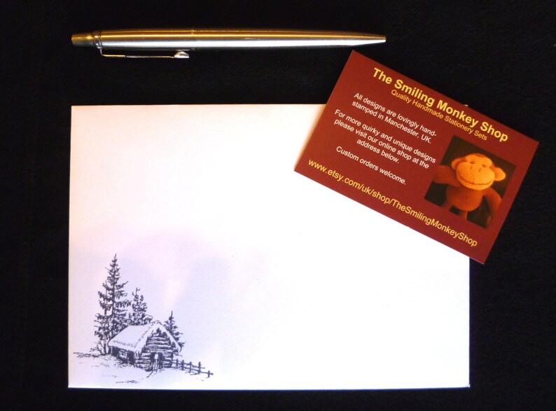 May include: A white card with a black and white illustration of a small cabin with trees. The card is next to a business card for The Smiling Monkey Shop, which features a smiling monkey illustration and the website address www.etsy.com/uk/shop/TheSmilingMonkeyShop.