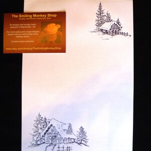 May include: A black and white illustration of a snowy cabin with a snow-covered roof. The cabin is surrounded by trees and a fence. The text "The Smiling Monkey Shop" is printed on the top of the image.