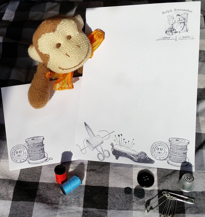 Puede incluir: Un mono de peluche mira por encima de una hoja de papel blanca con ilustraciones de costura. El papel presenta carretes de hilo, tijeras, un alfiletero y el texto "Dolly's Dressmaker London". Se ven botones, un dedal y alfileres de seguridad.