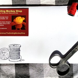 Puede incluir: Un juego de papeler&iacute;a blanco con una tarjeta de visita roja y naranja de The Smiling Monkey Shop. El juego incluye carretes de hilo rojo y azul, un dedal, tijeras y una imagen estampada de una bobina de hilo.