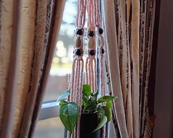 May include: A pink macrame plant hanger with wooden beads hangs in a window. A small green plant is in a black pot.