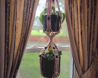 May include: A pink macrame plant hanger with two black pots and green plants hanging from a window.