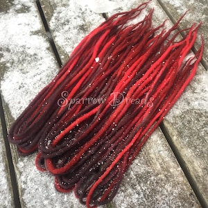 Puede incluir: Un paquete de rastas de lana roja y marrón oscuro, probablemente para extensiones de cabello. Las rastas están dispuestas en forma de abanico y están sobre una superficie de madera cubierta de nieve.