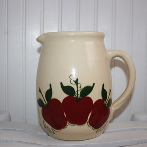 May include: Cream-colored ceramic pitcher with a handle and spout. The pitcher is decorated with three large red apples with green leaves and vines. A charming addition to any kitchen.