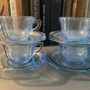 May include: Four light blue glass teacups and saucers are stacked in pairs. The cups and saucers have a floral pattern. The teacups have handles. The background includes books.