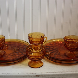 May include: Amber-colored vintage glassware set. The set includes footed cups, saucers, and plates. The glassware has a textured, geometric pattern. The set is displayed on a white surface against a white paneled background.
