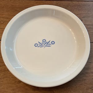 May include: Off-white pie plate with a blue floral design in the center. The plate has a wide rim and is made of ceramic. The floral design features stylized blue flowers and leaves. The plate is round and appears to be vintage.