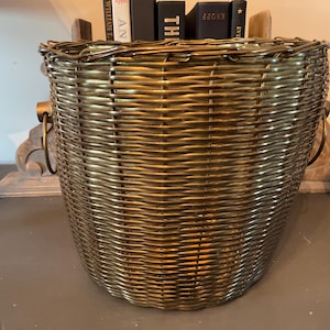 May include: A woven basket with a metallic gold finish. The basket has a rounded shape with a wide opening and two metal handles. The basket is filled with books. A decorative item is visible in the background.