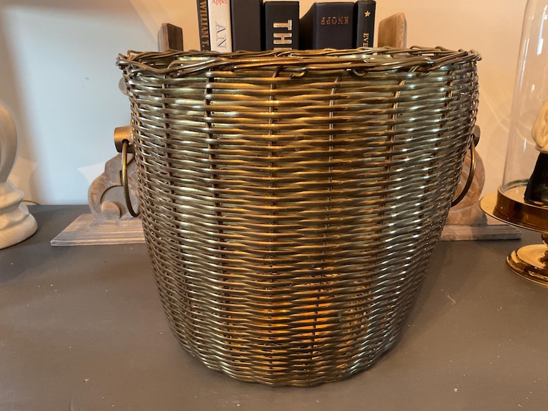 May include: A woven basket with a metallic gold finish. The basket has a rounded shape with a wide opening and two metal handles. The basket is filled with books. A decorative item is visible in the background.