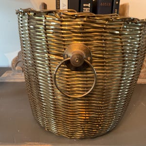 May include: A woven gold-colored basket with a brass handle. The basket has a rounded shape and is made of tightly woven material. The handle is a circular ring attached to a brass plate. The basket is likely used for storage or decoration.