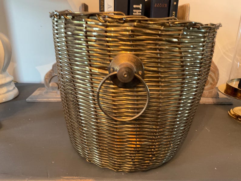May include: A woven gold-colored basket with a brass handle. The basket has a rounded shape and is made of tightly woven material. The handle is a circular ring attached to a brass plate. The basket is likely used for storage or decoration.