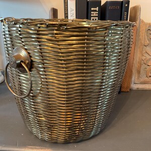 May include: A woven basket with a metallic gold finish. The basket has a rounded shape with a handle on one side. The basket is made of woven material and has a textured surface. The basket is empty.
