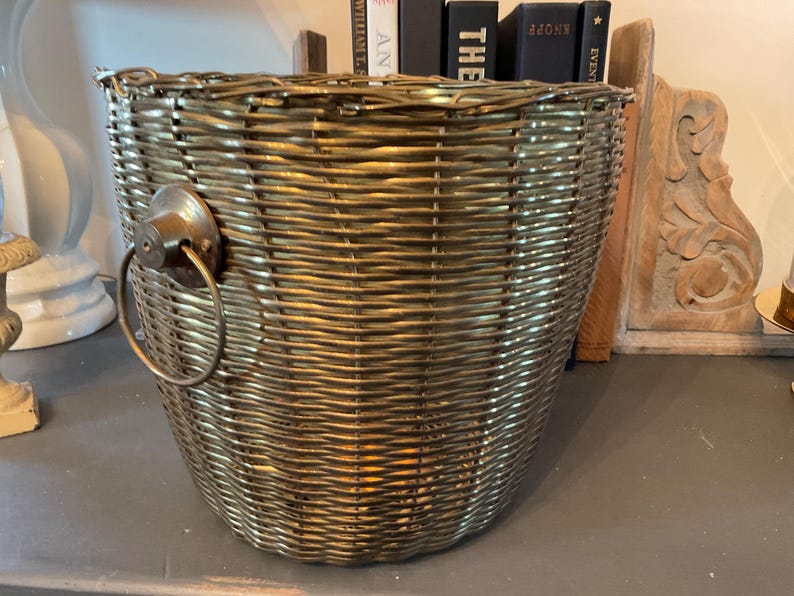 May include: A woven basket with a metallic gold finish. The basket has a rounded shape with a handle on one side. The basket is made of woven material and has a textured surface. The basket is empty.