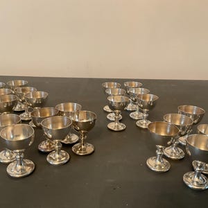 May include: A collection of small, silver-colored goblets with rounded bowls and pedestal bases. The goblets are arranged on a dark surface, with some grouped together and others spaced apart. The overall theme suggests a vintage or antique collection.