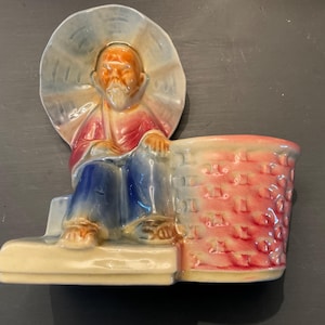 May include: A ceramic planter featuring a seated figure with a wide-brimmed hat. The figure wears a red shirt and blue pants, with a white beard. The planter has a brick-like texture and is painted in shades of red and pink.