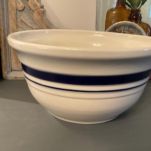 Puede incluir: Cuenco mezclador de cerámica color crema con dos rayas azul marino. El cuenco tiene forma redondeada con un borde ancho y un labio ligeramente ensanchado. Un artículo de cocina clásico.