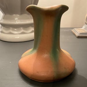 以下が含まれることがあります： フレアトップと丸みを帯びたベースのセラミック花瓶。花瓶は主にオレンジ色で、垂直の緑色のストライプがあります。花瓶は灰色の表面に置かれています。