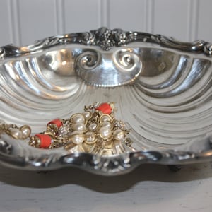 May include: A silver, shell-shaped dish with a decorative rim and a beaded necklace with orange and white beads inside.
