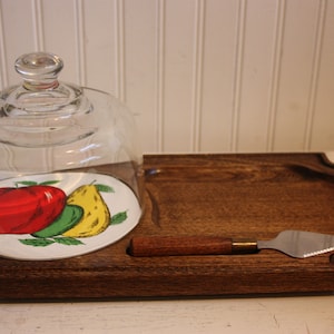May include: A wooden cheese board with a glass dome, a small plate with a painted design of fruit, and a small knife.