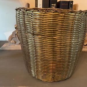 May include: A woven basket with a metallic gold finish. The basket has a rounded shape with a wide opening and two handles. The basket is made of a tightly woven material, and the gold finish gives it a vintage look.