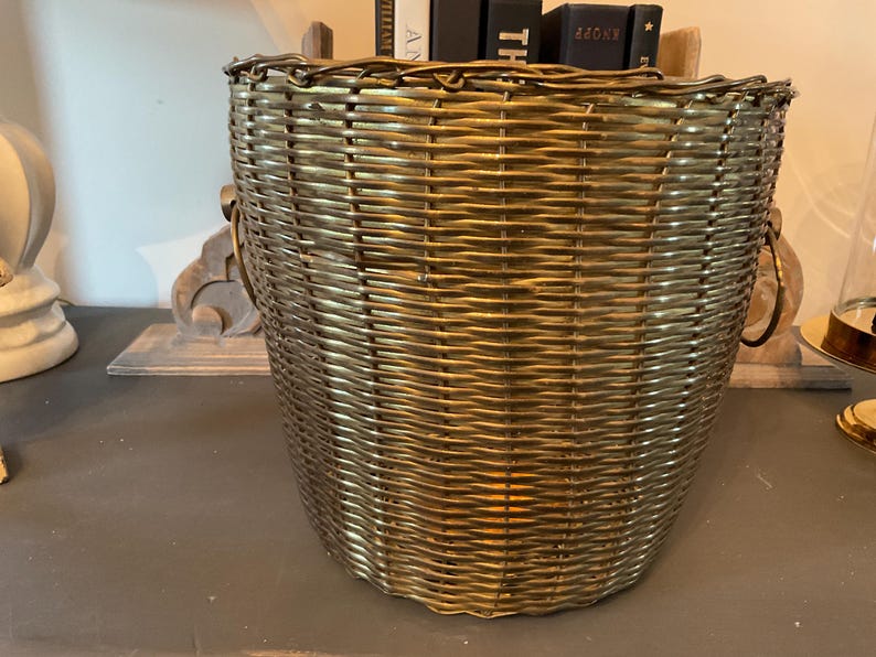 May include: A woven basket with a metallic gold finish. The basket has a rounded shape with a wide opening and two handles. The basket is made of a tightly woven material, and the gold finish gives it a vintage look.