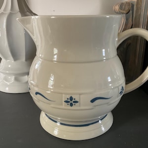 May include: Cream-colored ceramic pitcher with a rounded body and a handle. The pitcher features blue floral and wave designs around the base and body. A white decorative vase is in the background.