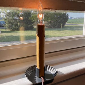 May include: An electric candle lamp featuring a tan, textured pillar with faux wax drips. The candle is set in a dark, circular, fluted metal base. A clear, flame-shaped light bulb is illuminated at the top, and a thin brown power cord extends downward.
